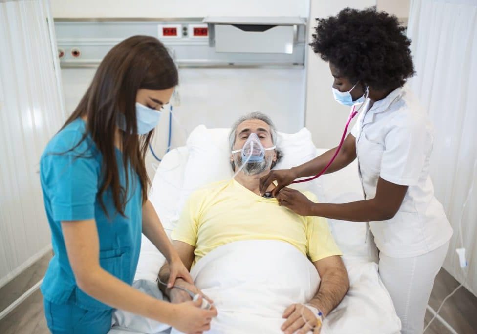 Young doctor and nurse wearing a surgical mask checking on a senior male patient wearing a positive pressure oxygen mask to aid breathing in a hospital bed during the Covid-19 pandemic